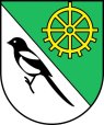 Im schräggeteilten Feld vorne in Silber eine schwarze Elster mit silberner Brust und silberner Armdecke, den Schnabel geöffnet, hinten in Grün eine zweigeteiltes goldenes Rad, die obere Hälfte als Wagenrad, die untere Hälfte als unterschlächtiges Mühlenrad ausgebildet.
